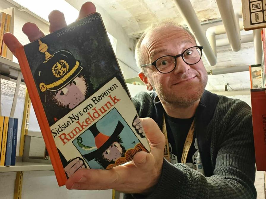 Formidler Hans Wendelboe Bøcher holder "Sidste nyt om Røveren Runkeldunk" frem foran sig, mellem magasinets reoler. 