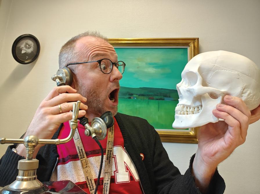 Kultur- og historieformidler Hans Wendelboe Bøcher i rød t-shirt sidder med et menneskekranium i den ene hånd og et gammelt telefonrør i den anden. Han er overrasket ud. I baggrunden hænger et grønt maleri og et portrtæt af N.F.S. Grundtvig. i 